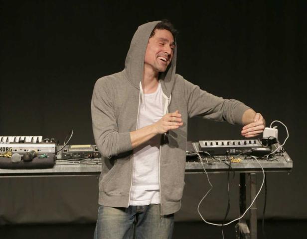 Ein Mann mit Kapuzenpullover steht lächelnd vor einem Tisch mit Synthesizern.