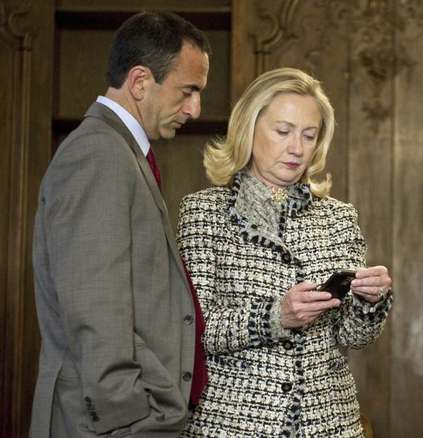 Hillary Clinton schaut auf ihr Smartphone, während ein Mann neben ihr steht.