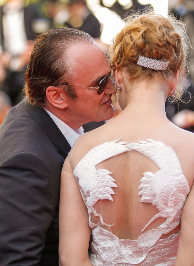 Quentin Tarantino flüstert einer Frau in einem weißen Kleid etwas ins Ohr.