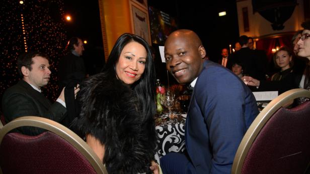 Eine Frau mit einer Pelzstola und ein Mann im blauen Anzug sitzen an einem Tisch bei einer Veranstaltung.