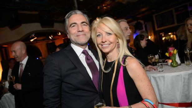 Ein Mann im Anzug und eine blonde Frau lächeln auf einer Party in die Kamera.