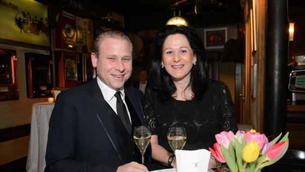 Ein Mann im Anzug und eine Frau in einem schwarzen Kleid sitzen an einem Tisch mit Sektgläsern und Tulpen.