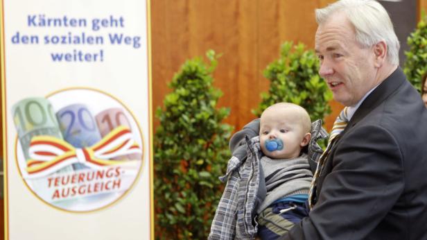 Ein Mann im Anzug hält ein Baby mit Schnuller im Arm; im Hintergrund ein Schild zum Teuerungsausgleich.