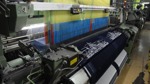 Ein Webstuhl produziert ein blaues Tuch mit einem weißen Stadtmotiv.