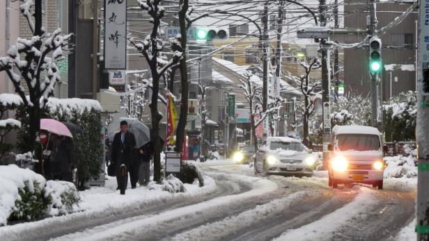 Eine verschneite Straße in einer japanischen Stadt mit Autos und Fußgängern.