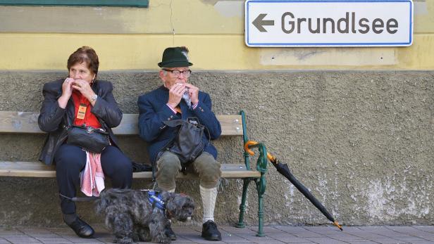 Ein älteres Paar isst auf einer Bank in der Nähe eines Schildes nach Grundlsee, mit einem Hund dabei.