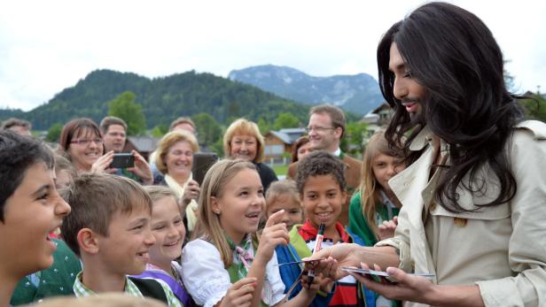 Conchita Wurst gibt Autogramme an eine Gruppe Kinder vor einer Bergkulisse.