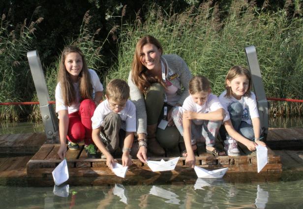Eine Frau und vier Kinder lassen Papierboote in einem See schwimmen.