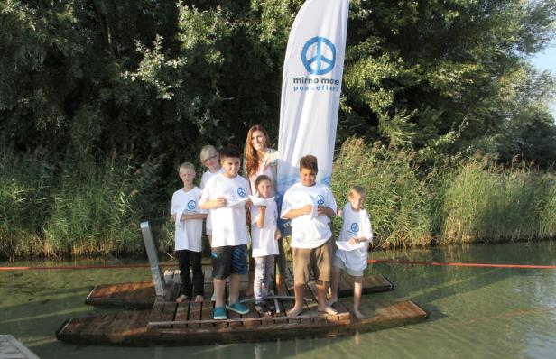 Eine Gruppe Kinder und eine Frau stehen auf einem Steg im Wasser, mit einem Friedensbanner im Hintergrund.