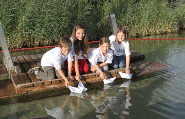 Vier Kinder lassen Papierboote von einem Holzsteg ins Wasser gleiten.