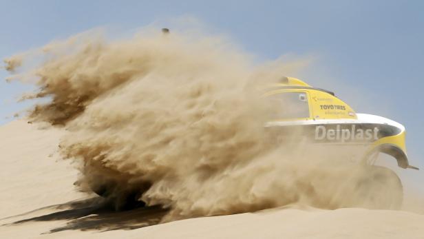 Ein gelb-weisses Rennauto fährt durch eine Sanddüne und wirbelt Staub auf.