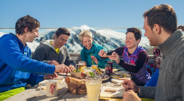 Eine Gruppe von Freunden isst und trinkt auf einer Terrasse mit Blick auf schneebedeckte Berge.