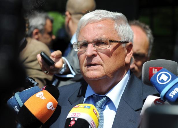 Ein Mann mit Brille wird von Reportern mit Mikrofonen umringt.