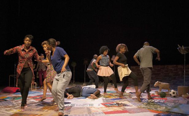 Eine Gruppe von barfüßigen Darstellern tanzt auf einer Bühne mit einem patchworkartigen Boden.