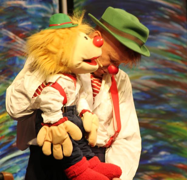 Ein Mann im Clownskostüm mit roter Nase und Hut hält eine Handpuppe.
