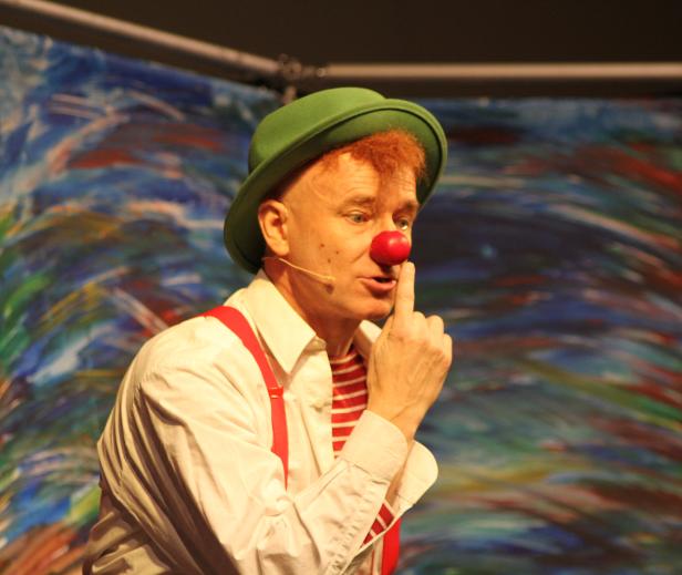Ein Clown mit roter Nase und grünem Hut deutet mit dem Finger nach oben.