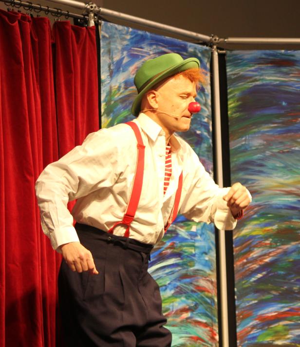Ein Clown mit roter Nase, grünem Hut und Hosenträgern auf einer Bühne.