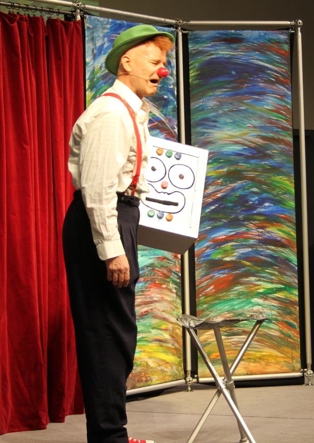 Ein Clown mit roter Nase und grünem Hut steht auf einer Bühne.