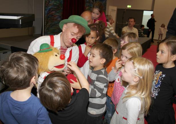 Ein Clown mit roter Nase und grünem Hut umringt von Kindern.