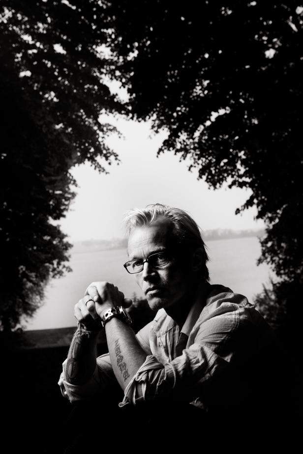 Ein Mann mit Brille und Tätowierungen sitzt vor einer Landschaft.