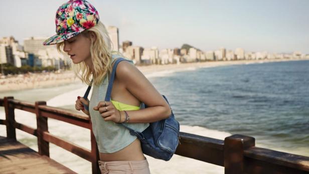 Eine junge Frau mit Blumenhut und Rucksack steht an einem Strand in Rio de Janeiro.