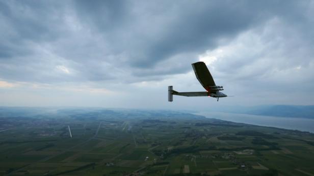 Ein Solarflugzeug fliegt über eine grüne Landschaft unter einem bewölkten Himmel.