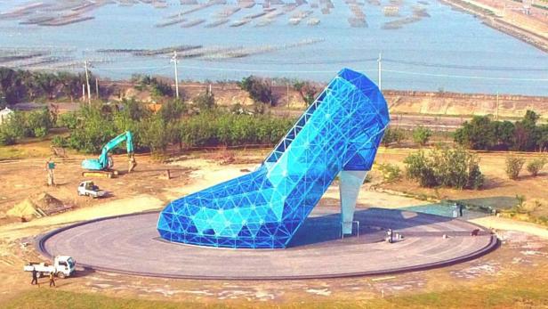 Eine riesige, blaue High-Heel-Skulptur steht auf einem runden Platz in Küstennähe.