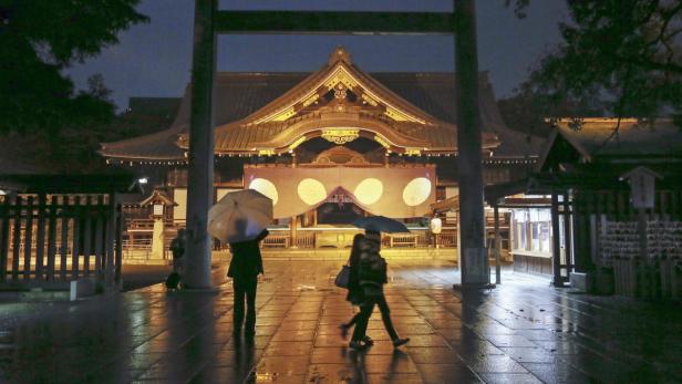 Menschen mit Regenschirmen vor einem beleuchteten japanischen Schrein bei Nacht.