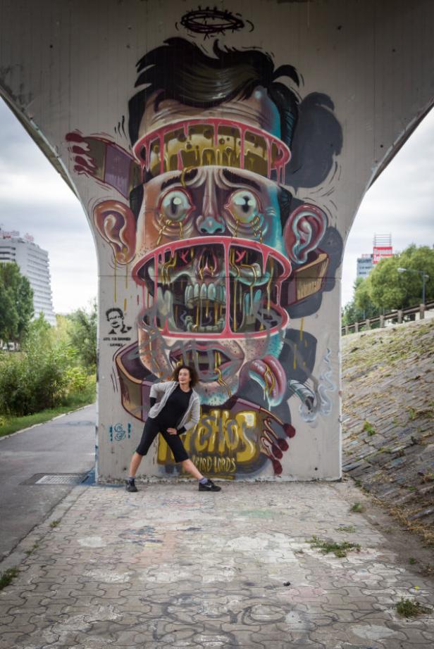 Eine Frau posiert vor einem Graffiti mit einem aufgeschnittenen Gesicht unter einer Brücke.