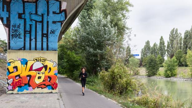 Eine Frau joggt auf einem Weg entlang eines Flusses, neben Graffitiwänden.