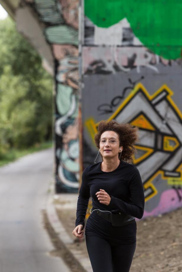 Eine Frau mit Kopfhörern joggt an einer mit Graffiti bemalten Wand entlang.