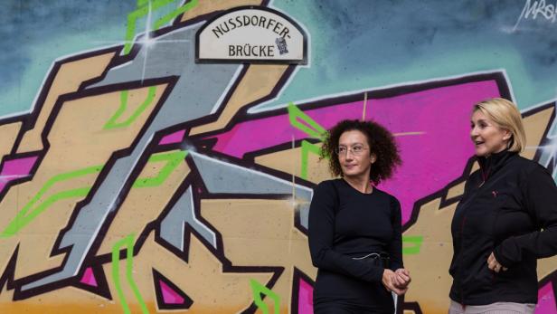 Zwei Frauen stehen vor einer Wand mit Graffiti und einem Schild „Nussdorfer-Brücke“.