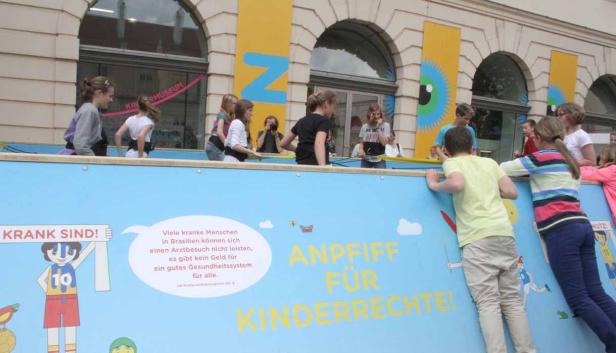 Eine Gruppe Kinder steht vor einer blauen Wand mit der Aufschrift „Anpfiff für Kinderrechte!“.