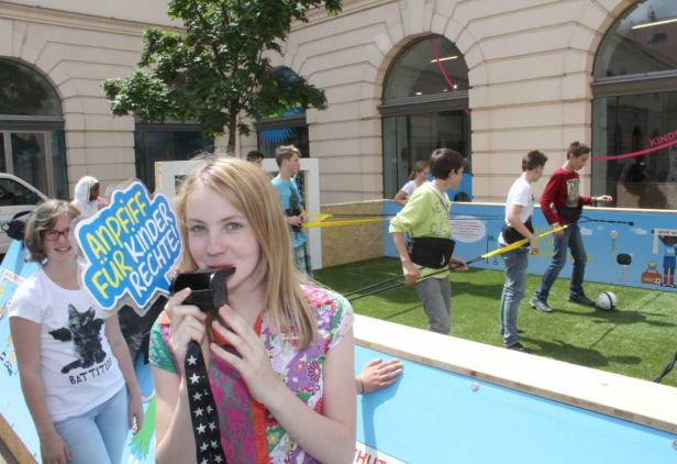 Ein Mädchen bläst in eine Trillerpfeife, während im Hintergrund Kinder Fußball spielen und ein Schild „Andiff für Kinder Rechte!“ zu sehen ist.
