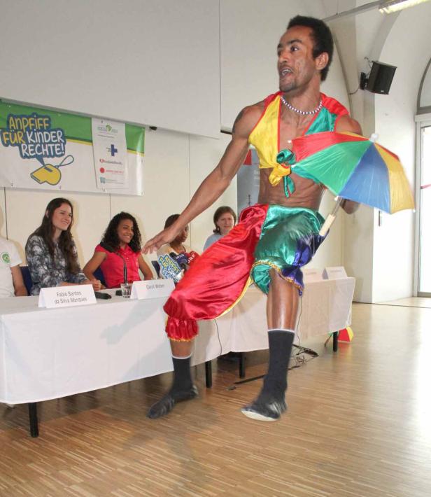 Ein Mann in farbenfroher Kleidung tanzt vor einem Tisch mit Personen und einem Banner für Kinderrechte.