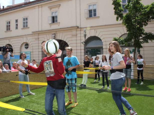 Eine Gruppe von Kindern spielt ein Spiel mit Gummibändern und einem Ball im Freien.