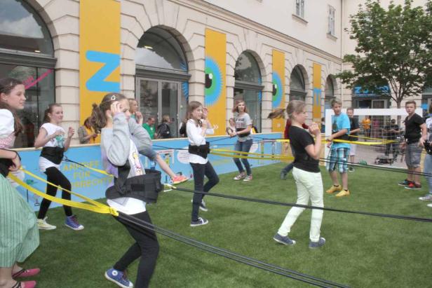 Eine Gruppe Kinder spielt mit Gummibändern auf einer Wiese vor einem Gebäude.