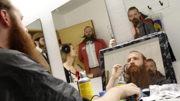 Ein Mann formt seinen Bart vor einem Spiegel, während andere Personen im Hintergrund zu sehen sind.