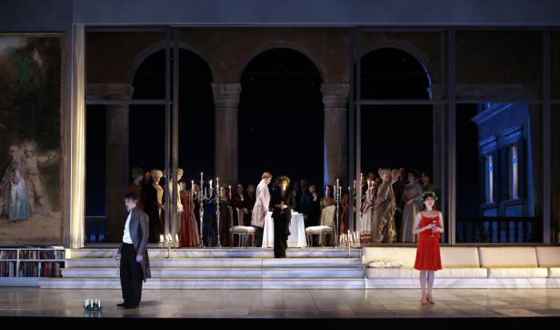 Eine Theaterszene mit Schauspielern auf einer Bühne vor einem gemalten Hintergrund.