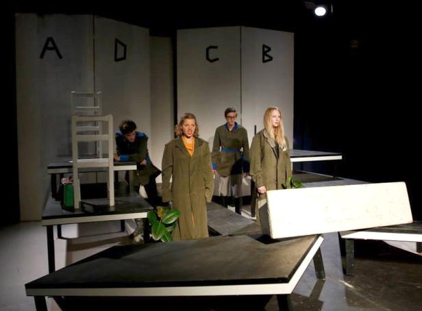 Vier Schauspieler stehen auf einer Bühne mit minimalistischem Bühnenbild und Buchstaben im Hintergrund.