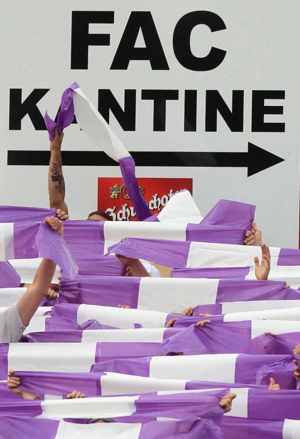 Fans schwenken lila-weiße Fahnen vor einem Schild mit der Aufschrift „FAC Kantine“.