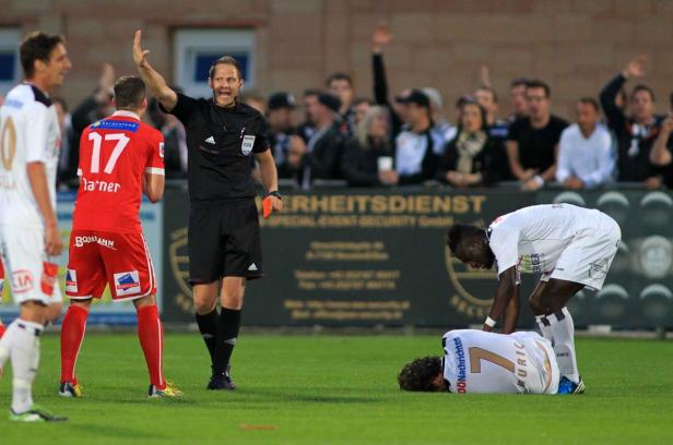 Ein Fußballschiedsrichter zeigt einem Spieler die Rote Karte, während ein anderer am Boden liegt.