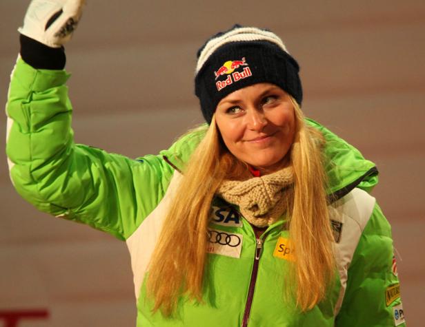 Lindsey Vonn mit Mütze und grüner Jacke lächelt und hebt die Hand.