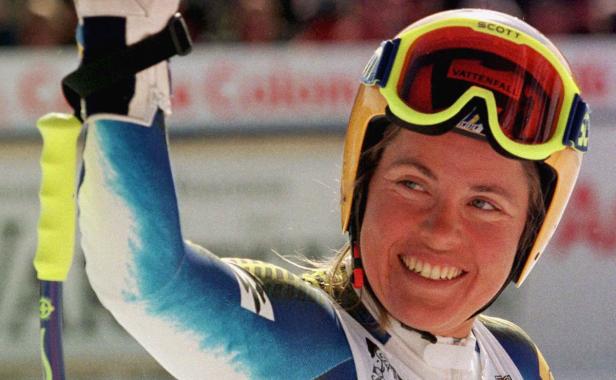 Eine lächelnde Skirennläuferin mit gelbem Helm und Skibrille hebt den Arm.