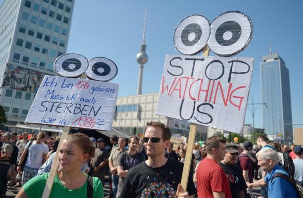 Menschen demonstrieren mit Schildern, auf denen „Stop Watching Us“ und „Ich bin 1984 geboren. Muss ich auch sterben wie in 1984?“ steht.