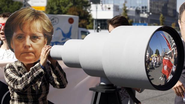 Eine Person mit einer Angela-Merkel-Maske blickt durch ein übergroßes Fernrohr auf eine Demonstration.