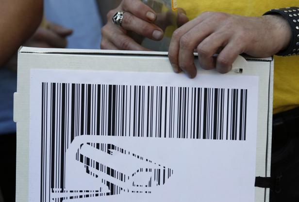 Eine Person hält ein Schild mit einem Barcode und einer stilisierten Überwachungskamera.