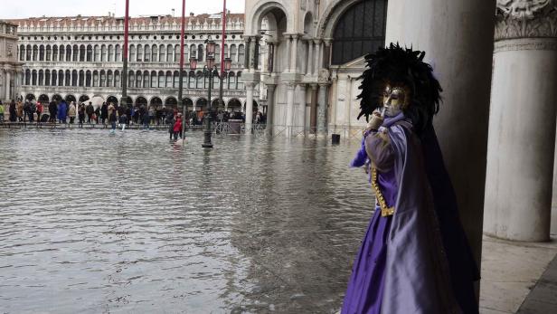 Eine Person in venezianischer Maske und Kostüm steht vor dem überfluteten Markusplatz.