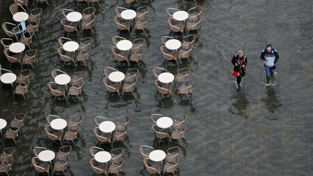 Zwei Personen waten durch das überflutete Venedig, Tische und Stühle eines Cafés stehen im Wasser.