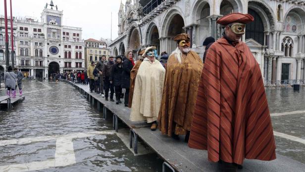 Menschen in Venedig während Hochwasser, einige mit Masken und Kostümen, gehen auf einem erhöhten Steg.
